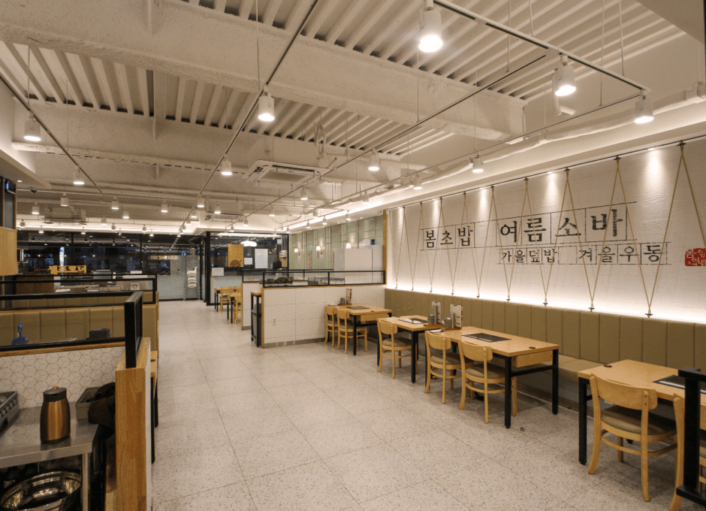 전주 식당 인테리어 추천 사례로 손꼽히는 일식 매장의 내추럴 우드 톤 테이블 가구 배치와 감각적인 벽면 간접 조명 시공 완료 사진 통로